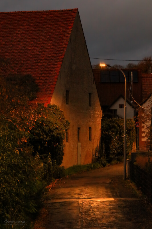 Evening photo of an old alley leading into a German village, under a heavily clouded gray sky. On the left is some foliage, then the large red tile roof and facade of a huge stone barn. The peak of the roof is three tall stories high, and it slopes down to the ground floor. The barn, and the moist street, are illuminated by the soft golden glow of a streetlamp.