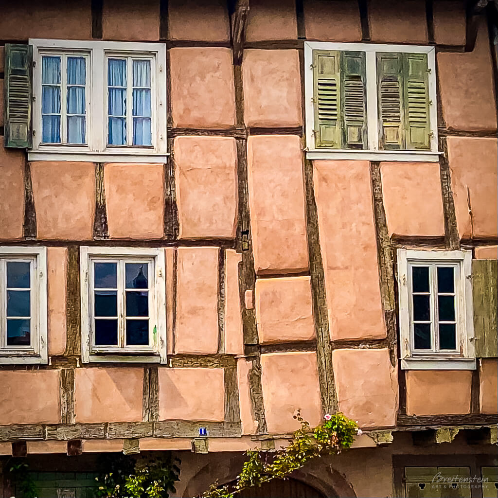 Photo of a wall of a half-timbered house, painted a salmon color, with five windows (or 7 if you’re picky). Some windows have old-style shutters, and the timber beams are quite uneven, demonstrating that the house is very old. 