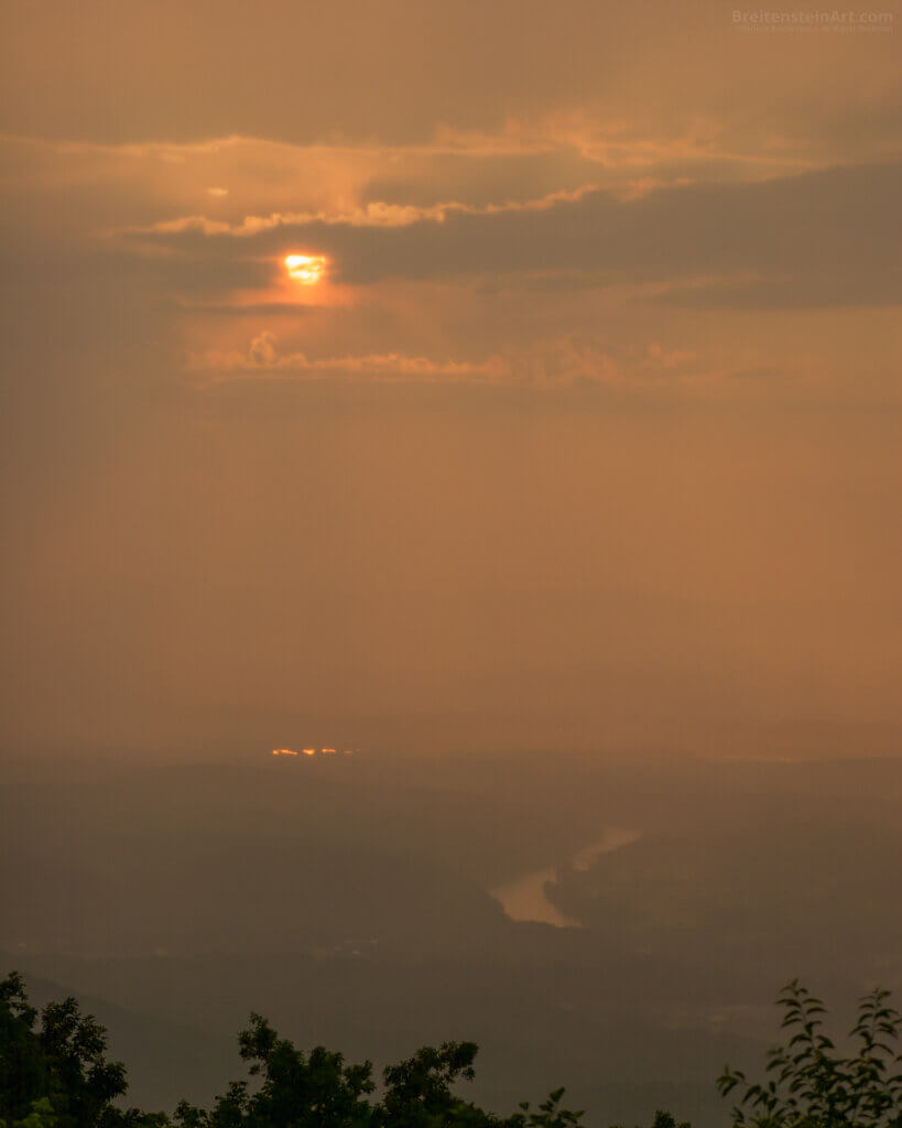 Photograph of a very hazy orange sunset, over an indistinct landscape that includes the Shenandoah River. There are a few branches of trees at the very bottom.