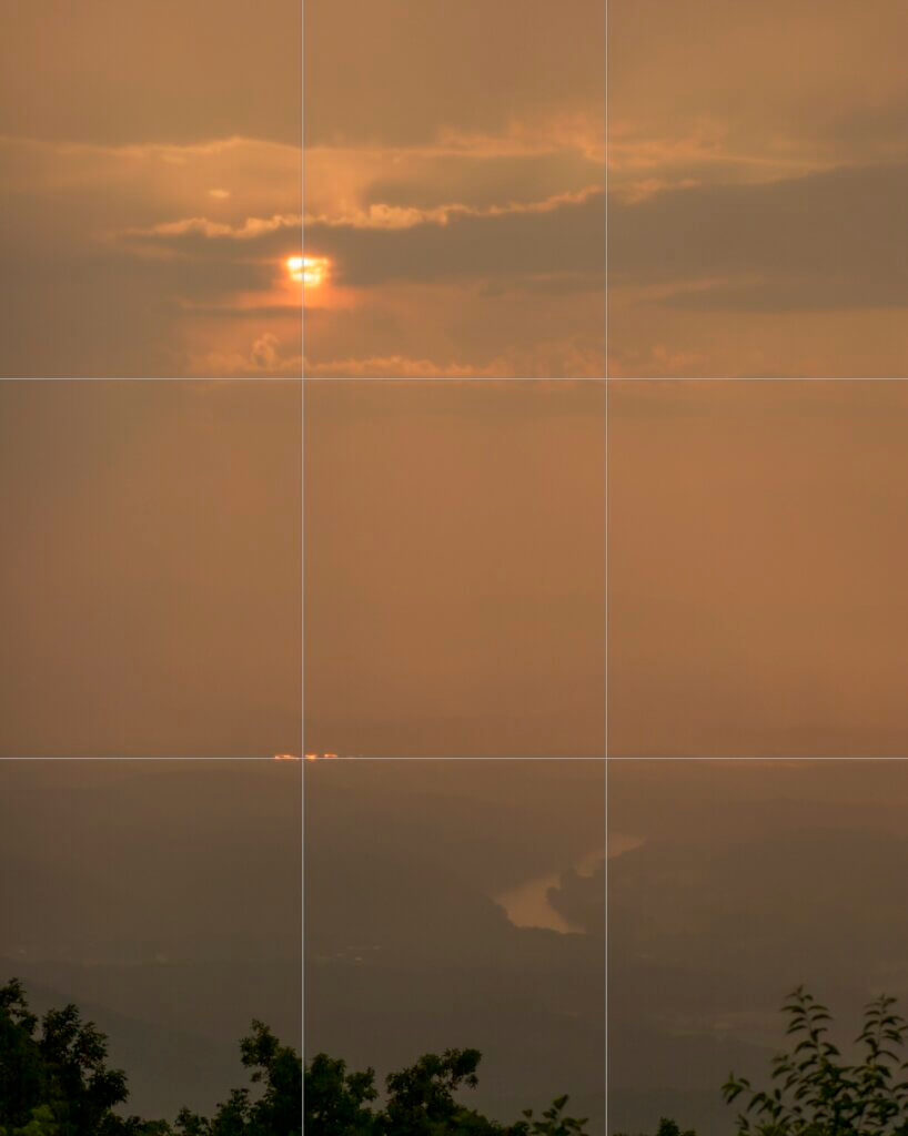 Photograph of a very hazy orange sunset, over an indistinct landscape that includes the Shenandoah River. There are a few branches of trees at the very bottom. Overlaid on the photo are two horizontal and two vertical lines, breaking the image into an even 3x3 grid.