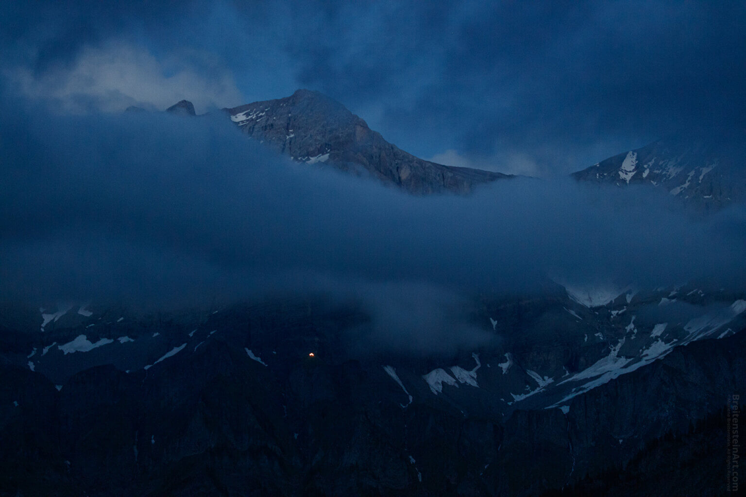 Photograph near dark, of snow-streaked mountain peaks, partly obscured by a band of clouds, under a cloudy sky. The scene is a gamut of muted blues, and there is a very tiny yellow light, the illuminated peak of a house on the mountainside.