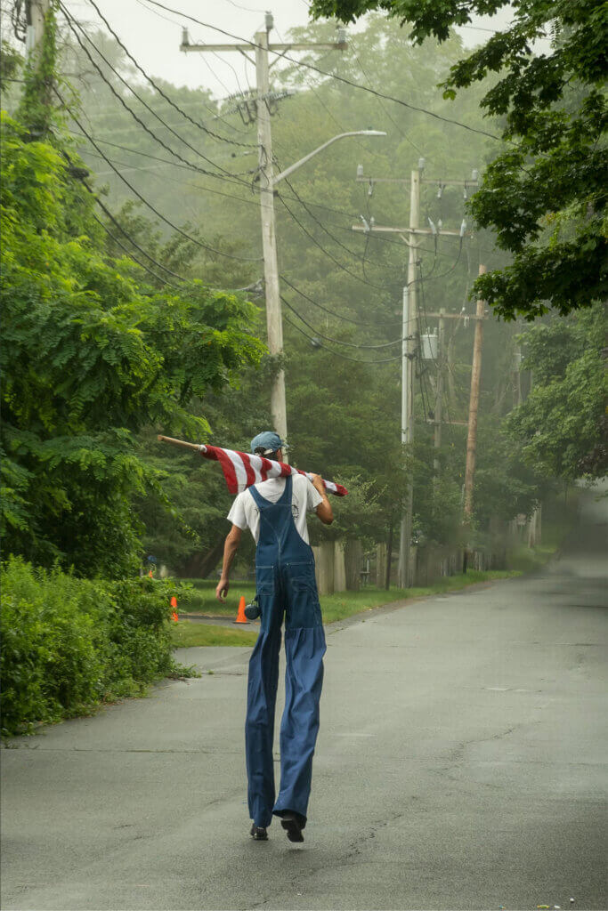 Photo of a man in stilts, wearing very long overalls, walking away from the viewer, down a misty country road. The man holds an American flag, rolled on its pole, over his shoulder.