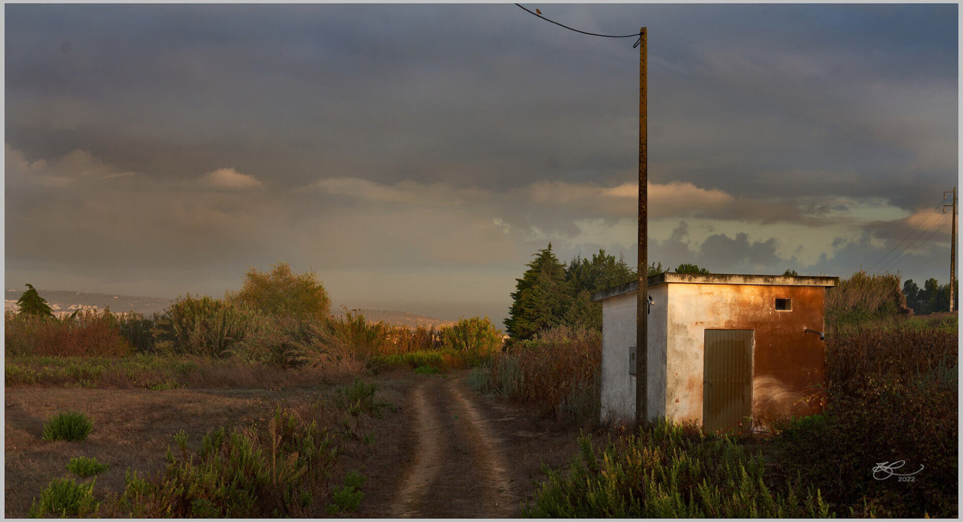 Photograph of an unpaved rural road among brush, under muted, post-sunset clouds. Next to the road is an electric pole and a small, simple building, painted white and rust-red.