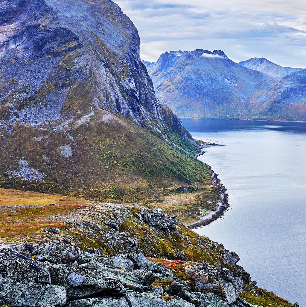 Photo of a gorgeously colorful, rugged mountain landscape, bisected by a large body of water. Almost imperceptible on the shoreline, are a few tiny buildings, hinting at the enormous scale of the landscape.