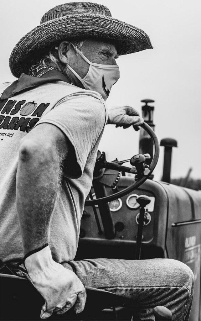 Black and white photo of a man in a cowboy hat, farm clothes, a bandana, and work gloves. He is wearing a cloth mask and sitting on a tractor, looking off into the distance.