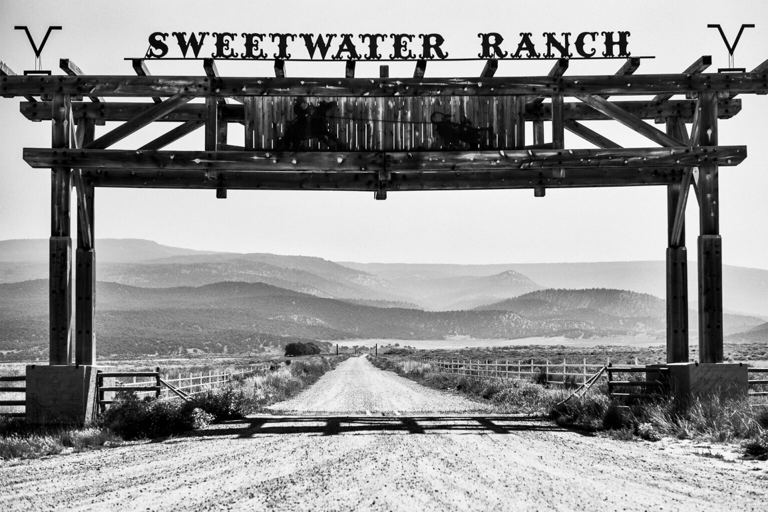 Black and white photo of the entrance and sign to Sweetwater Ranch, over a road leading to a wide, low mountain vista in the distance.