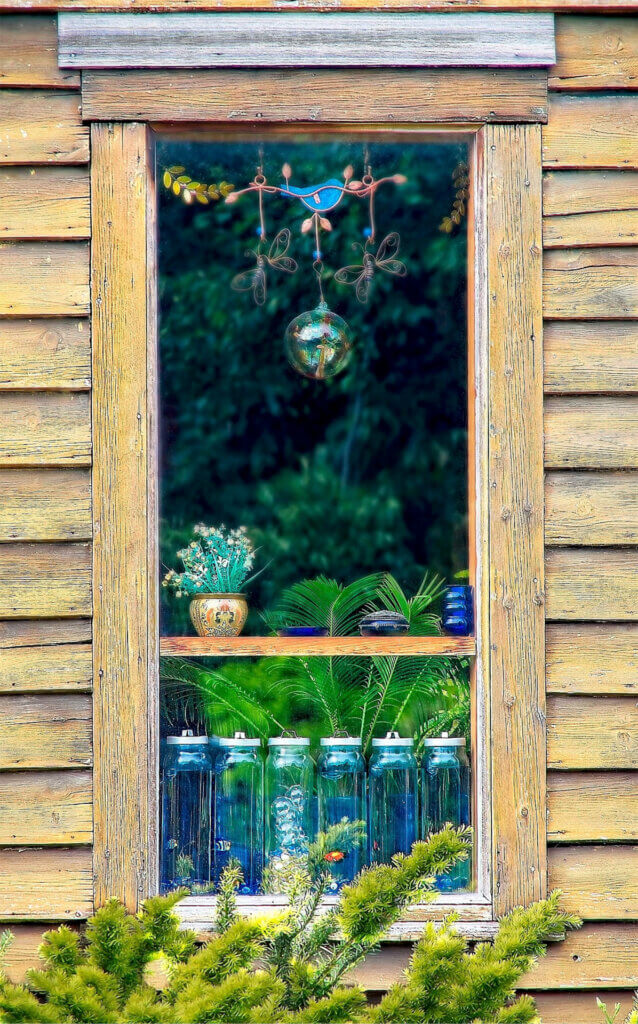 Photo of the window of a rural home, its frame and the siding a faded yellow, through which we see a decorative glass hanging, six glass storage bottles, small bowls and houseplants.