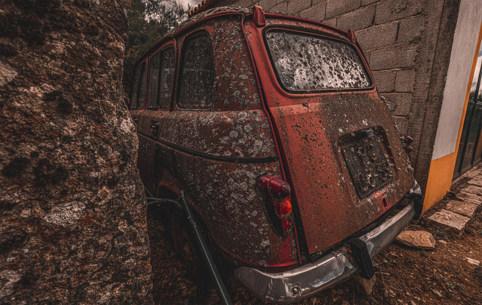 Photo of an old European car that has sat abandoned for a long time. It is between an earthen mound and a cinder block building. The car is red, but it is largely covered with dirt, mold and large expanses of lichen.