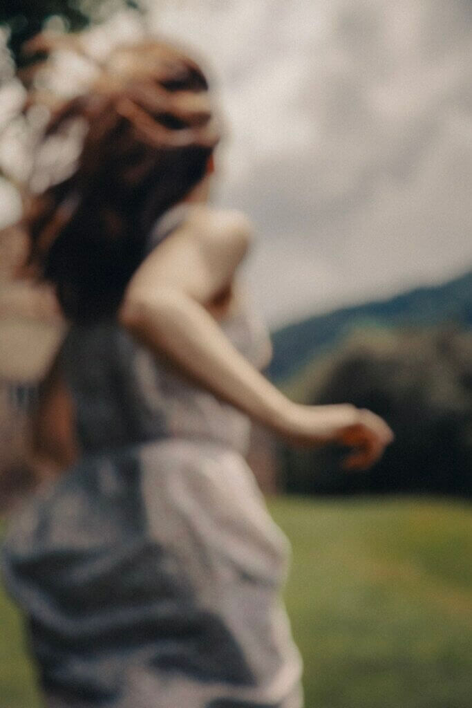 Photograph in soft focus, of a woman in a dress, running in a field. She is seen from the back from the thigh up, her hair streaming out, and it appears she has just passed the photographer.