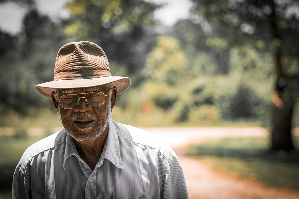 Photo of a man wearing wire-frame glasses, under a banded, straw, Stetson-style hat, and a thin-striped button down shirt, open at the collar. His is older, with a very short, gray beard and gray eyebrows. The background, of a curving road and trees, is softly blurred.