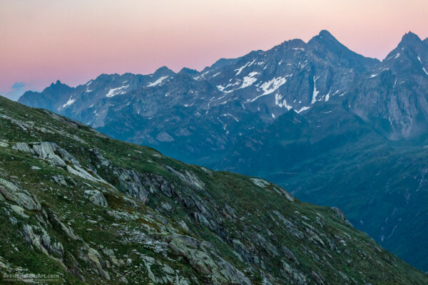 Photograph of a mountainous vista. In the foreground is a rocky hillside of many textures and colors, in shades of grays and greens. In the back we see a snow-speckled mountain range, consisting of many crags and peaks, in shades of blue, green and violet. The sky above is a soft but bright gradation of yellow to pink.